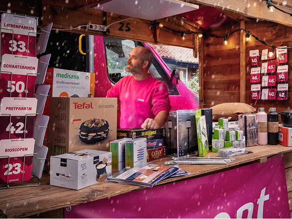 Man in een roze trui staat in een houten kraam vol cadeauproducten en kortingskaarten, terwijl het sneeuwt.