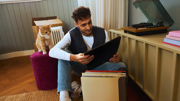 Iemand bladert door een stapel lp’s, met een nieuwsgierige kat naast zich.